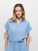 A woman with light brown hair wears the Maddie Blouse In Linen—relaxed fit, garment-dyed, in light blue—tucked into matching drawstring pants. She smiles softly at the camera against a plain white background.