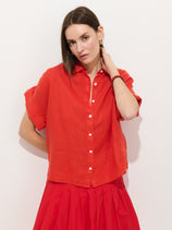 A woman with straight brown hair wears the Maddie Blouse In Linen— a relaxed fit, short-sleeved red button-up with shell buttons—paired with a matching red skirt, standing against a white wall and touching her neck.