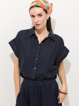 A woman wears the Maddie Blouse In Linen, garment-dyed in dark navy with a relaxed fit. She pairs it with a colorful headscarf and red beaded bracelet, posing against a light background with her hands in her pockets.