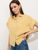 A woman wearing the Maddie Blouse In Linen, a relaxed fit garment-dyed yellow shirt, with blue jeans stands against a white background, one hand on her hip and a neutral expression.