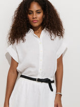 A woman with curly hair wears the Maddie Blouse In Linen—a relaxed fit, 100% linen short-sleeve button-up—paired with a matching white skirt and black braided belt, standing against a plain white background.
