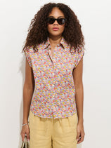Wearing dark sunglasses, a person with curly hair models the Maddie Blouse in Liberty Cotton— a sleeveless button-up shirt featuring pink, yellow, and orange floral patterns—paired with light yellow shorts and holding a bag against a white background.