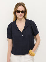 A woman with light brown hair, wearing sunglasses and the Madeleine Shirt in Linen styled with white pants, stands against a plain white wall. She wears a yellow scrunchie on her wrist.