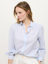 A woman with light brown hair wears the Hannah Striped Shirt In Cotton Voile, featuring shell buttons and slim vertical stripes, paired with white pants. She stands against a plain light background, gazing slightly to her left.