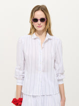 A woman in pink sunglasses wears the Hannah Striped Shirt In Cotton Voile, a white blouse with thin vertical stripes, standing against a plain white background and holding a red flower in her left hand.