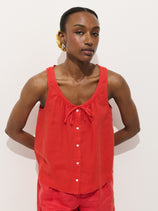 A woman stands against a white background, wearing the Sidney Top In Linen—a sleeveless red button-up with a bow neckline—paired with matching relaxed fit red linen shorts. Her hands are behind her back as she looks calmly at the camera.