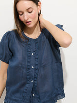 A woman with light skin and straight brown hair wears the Brittany Top In Ramie, a blue short-sleeve blouse with ruffle and button details. She looks down, touching her neck, against a plain light background.