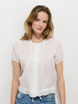 A woman with light brown hair wears the Brittany Top In Ramie, a short-sleeved white blouse with ruffle details, paired with blue jeans. She stands against a plain white background and gazes at the camera with a soft smile.