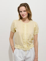 A woman with light brown hair wears the Brittany Top In Ramie, a pale yellow, short-sleeved, ruffled blouse made from 100% ramie fabric, paired with white pants, standing against a plain white background and smiling slightly.