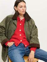 A woman in the Chiltern Street Jacket In Waxed Cotton with brown cuffs and collar, a red knit cardigan with white buttons, and blue jeans sits on a wooden chair against a plain background.
