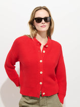 A person wearing dark sunglasses, olive green pants, and the Nico Cardigan in Cotton with corozo buttons stands against a plain white background.