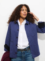 A woman with curly hair wears the Britt Work Jacket In Herringbone in blue over a white shirt and jeans, holding a maroon sweater and touching her hair, standing against a plain white background.