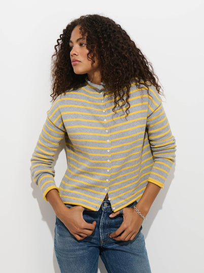 A person wearing the Taylor Striped Cardigan in Cotton Cashmere—featuring gray and yellow stripes—paired with blue jeans stands against a white background, looking to the side with one hand in their pocket.