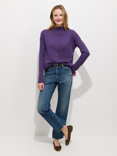 Standing against a white wall, the person is wearing the Taylor Cardigan in Cotton Cashmere—a purple oversized boxy fit long-sleeve top detailed with Troca shell buttons—paired with blue jeans, a black belt, and leopard-print flats. They have one hand on their hip and are smiling.