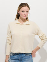 A woman with light skin and straight, light brown hair stands against a white background, wearing the cream-colored Taylor Cardigan in Cashmere and blue jeans, one hand on her hip and a neutral expression.