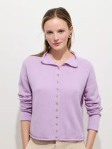 A woman with light brown hair wears the Taylor Cardigan In Cashmere, an oversized light purple women's knitwear button-up sweater, paired with beige pants, and stands against a white background looking at the camera with a slight smile.