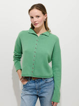 A woman models the Taylor Cardigan In Cashmere in light green, featuring an oversized fit, paired with blue jeans. She stands against a plain white background, one hand in her pocket and a slight smile on her face.