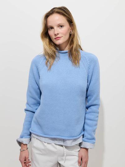 A woman with light brown hair stands against a white background, calmly looking at the camera while wearing the Eastport Rollneck In Cotton over a striped shirt and white pants.
