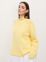 Woman with long brown hair wears the Eastport Rollneck in 100% cotton, yellow, paired with a white skirt. She stands against a plain white background, hands in pockets, gazing slightly upward.