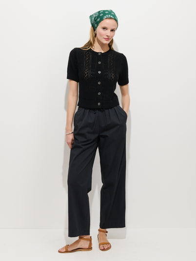 A woman stands against a white wall, wearing a green bandana, black short-sleeve buttoned knit top, the Hudson Pant in cotton twill, and brown sandals. Her hands are in her pockets and she is smiling slightly.
