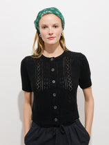 A woman stands against a white background, wearing a green bandana and the Elle Pointelle Cardigan in linen cotton—a black, short-sleeved piece with subtle pointelle stitching. She has her hands in her pockets and looks at the camera.