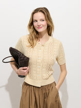 A woman wears the Elle Pointelle Cardigan in Linen Cotton and a brown skirt, posing against a white background with a woven dark brown bag, one hand in her pocket and a soft smile.