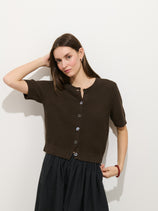 A woman with straight brown hair wears the Elle Cardigan In Linen Cotton, a hand-finished, dark brown short-sleeved piece with Troca shell buttons, paired with a black skirt. She stands against a white wall, one arm raised and hand touching her head.