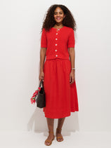 A woman with curly hair wears a bright red Maddie Skirt in Linen with a matching linen button-up top, holding a black bag with a red scarf and wearing brown sandals, standing against a plain white background.