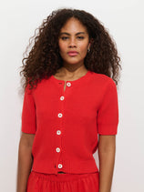 A woman with curly hair stands against a white background, wearing the bright red Elle Cardigan In Linen Cotton with Troca shell buttons and matching red bottoms. She looks directly at the camera with a neutral expression.