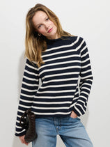 A woman with light brown hair wears the Eastport Striped Rollneck In Cotton and blue jeans, holding a dark woven bag. She stands against a white background, posing with one hand in her pocket and her head slightly tilted.