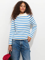 A person with curly hair wears the Eastport Striped Rollneck In Cotton sweater and loose blue jeans, holding a red item, standing against a plain white background.