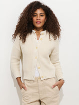 A woman with curly hair wears the Nico Cardigan in Cotton over a white top and beige pants, standing against a white background with her hands in her pockets, looking confidently at the camera.