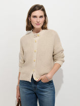 A person stands against a white background, wearing the Nico Cardigan In Cotton—a textured beige knit with large corozo buttons—paired with blue jeans. With shoulder-length brown hair and a relaxed expression, one hand in their pocket, they effortlessly showcase timeless women's knitwear style.
