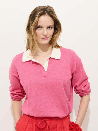 A woman with light brown hair wears the Bond St Polo In Cotton Cashmere in pink with a white collar and red pants, standing against a plain white background and looking at the camera with a slight smile.