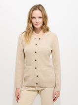 A woman with light brown hair wears the Bordeaux Cardigan in Cashmere, a beige two-ply cashmere piece featuring genuine horn buttons and front pockets, paired with beige pants. She stands against a plain white background, facing forward and smiling slightly.