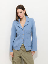 A woman with straight brown hair and a slight smile models the London Sweater Jacket in Cotton Cashmere—a light blue milano stitch style with four front pockets, brass crest buttons, and olive green cargo pants, against a white background.