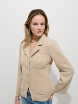 A woman with light brown hair wears the London Sweater Jacket In Cotton Cashmere, featuring a beige hue and large pockets, paired with blue jeans. She stands against a white background, looking at the camera with a slight smile.