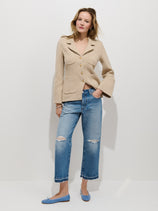 A woman with light, wavy hair poses confidently against a white background, wearing a beige button-up jacket, Drew Jean in a relaxed boyish fit, and light blue flats with one hand in her pocket.