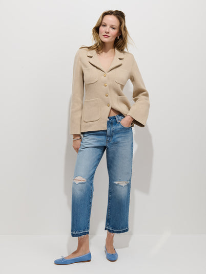A woman with light, wavy hair poses confidently against a white background, wearing a beige button-up jacket, Drew Jean in a relaxed boyish fit, and light blue flats with one hand in her pocket.