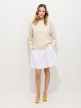 A woman with light brown hair smiles softly at the camera, hands in her pockets. She wears a cream cable-knit sweater, yellow shoes, and the Pippa Skirt in Cotton against a plain white background.