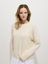 A woman with wavy, light brown hair wears the cream-colored Charlie Cable Sweater In Cashmere and a white skirt, standing against a plain white background and looking at the camera with a neutral expression.