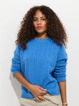 A person with curly brown hair wears the Charlie Cable Sweater In Cashmere in bright blue with beige pants, standing against a plain white background and looking calmly at the camera.