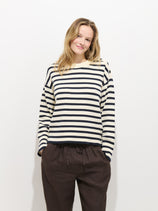 A person with shoulder-length blonde hair, smiling against a plain white background, wears the Normandy Striped Sweater in Cotton—black and white, 100% cotton, 7-gauge knit—and dark, loose-fitting pants with hands in pockets.