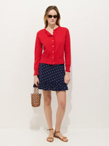 A woman stands against a plain white background wearing a red button-up cardigan, the Flora Skirt In Polka Dot with an elastic waistband and ruffle hem, brown sandals, sunglasses, and holding a brown woven handbag.