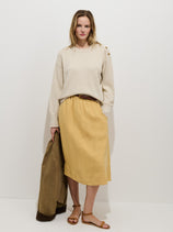 A woman with light brown hair stands against a white wall in a relaxed pose, wearing the Maddie Skirt In Linen—a cream 100% linen, garment-dyed skirt. She pairs it with a tan blouse, brown sandals, and holds a brown jacket.