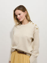 A woman with light brown hair stands against a white background, wearing the Normandy Sweater In Cotton with nautical shoulder buttons and a light tan skirt with a brown belt. She looks slightly to the side and smiles softly.