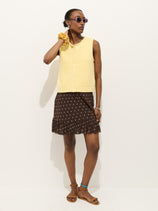 A woman poses against a white background wearing purple sunglasses, a sleeveless yellow top with buttons, the Flora Skirt In Polka Dot featuring an elastic waistband and ruffle hem, brown sandals, and a yellow scrunchie on her wrist.