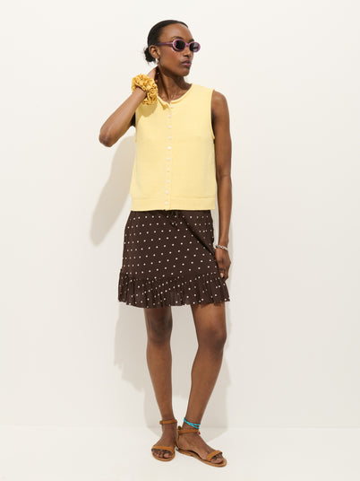 A woman poses against a white background wearing purple sunglasses, a sleeveless yellow top with buttons, the Flora Skirt In Polka Dot featuring an elastic waistband and ruffle hem, brown sandals, and a yellow scrunchie on her wrist.