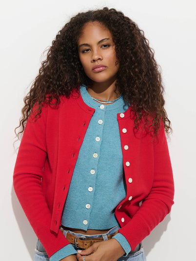 A person wearing a light blue Elizabeth Street Cardigan in Merino Wool layered over a bright red button-down sweater.