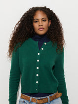 A person wears a forest green Elizabeth Street Cardigan in Merino Wool over a dark blue turtleneck, paired with blue jeans and a brown belt.